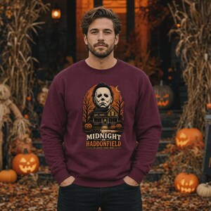 May include: A maroon long-sleeve sweatshirt featuring a graphic print of a house, a masked figure, and pumpkins. The text reads MIDNIGHT ON HADDONFIELD and HE ALWAYS COMES HOME. The design includes bare trees and a crescent moon against an orange background.