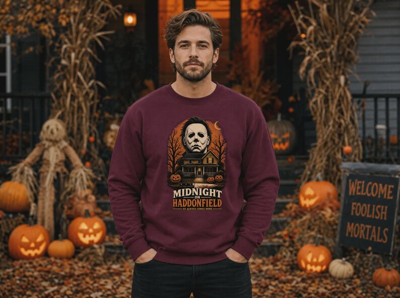May include: A maroon long-sleeve sweatshirt featuring a graphic print of a house, a masked figure, and pumpkins. The text reads MIDNIGHT ON HADDONFIELD and HE ALWAYS COMES HOME. The design includes bare trees and a crescent moon against an orange background.