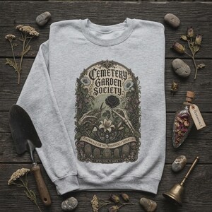 May include: A light grey crewneck sweatshirt featuring a detailed graphic print with the text Cemetery Garden Society, Perennials for the Permanently Departed, and Often Dusk to Dawn. The design includes skeletal hands, flowers, and tombstones.