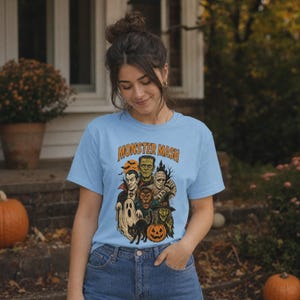 May include: A light blue short-sleeve t-shirt featuring a graphic print with the text MONSTER MASH in orange letters. The illustration depicts a group of classic Halloween characters including a vampire, Frankenstein, a mummy, a werewolf, a ghost, a witch, a black cat, and a jack-o'-lantern.