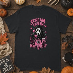 May include: A black short-sleeve t-shirt featuring a graphic print of a hooded figure in a white mask holding a pink rotary phone. The text reads "SCREAM QUEEN" and "DON'T HANG UP" in pink, surrounded by pink hearts, roses, and a drink cup.