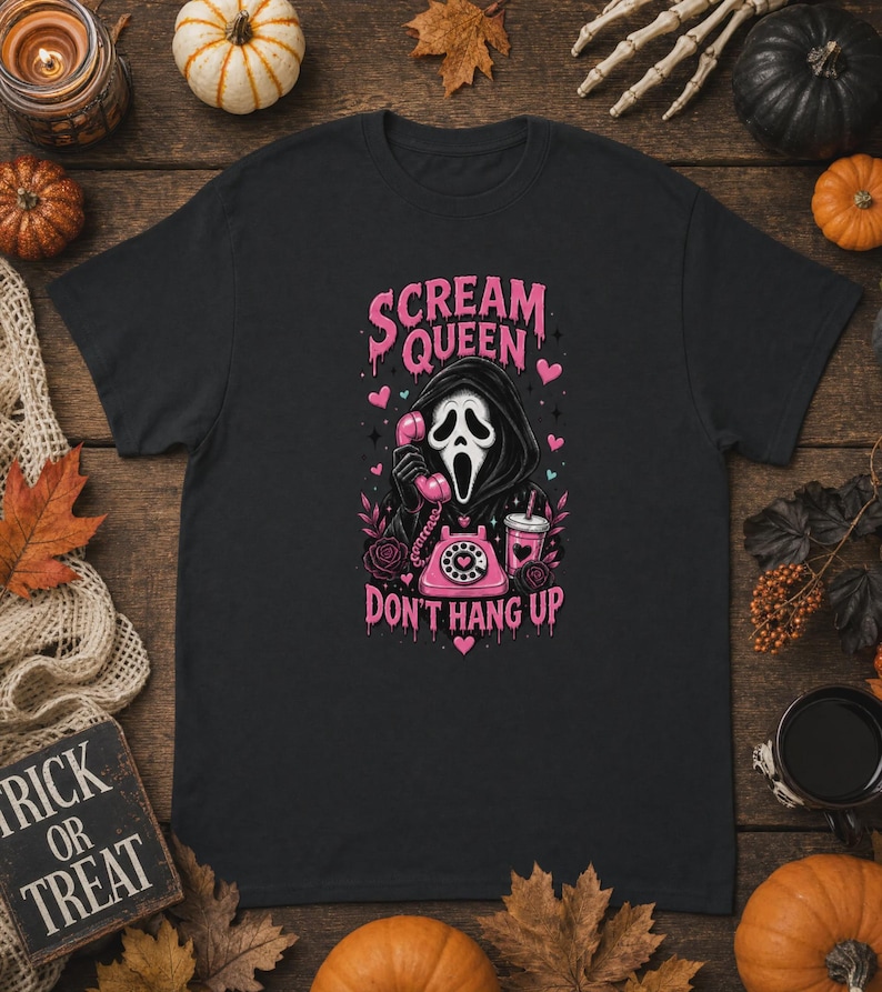 May include: A black short-sleeve t-shirt featuring a graphic print of a hooded figure in a white mask holding a pink rotary phone. The text reads "SCREAM QUEEN" and "DON'T HANG UP" in pink, surrounded by pink hearts, roses, and a drink cup.