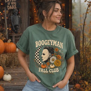 May include: A sage green short-sleeve t-shirt featuring a graphic print with the text BOOGEYMAN FALL CLUB. The design includes a profile illustration of a masked figure, a black and white checkered crescent moon, orange and white flowers, and pumpkins.