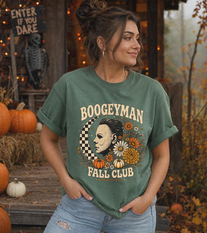 May include: A sage green short-sleeve t-shirt featuring a graphic print with the text BOOGEYMAN FALL CLUB. The design includes a profile illustration of a masked figure, a black and white checkered crescent moon, orange and white flowers, and pumpkins.