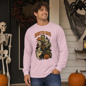 May include: A light pink long-sleeve shirt featuring a graphic print with the text MONSTER MASH. The design includes illustrations of Frankenstein, a vampire, a mummy, a werewolf, a ghost, a witch, a black cat, and a pumpkin in orange and green.