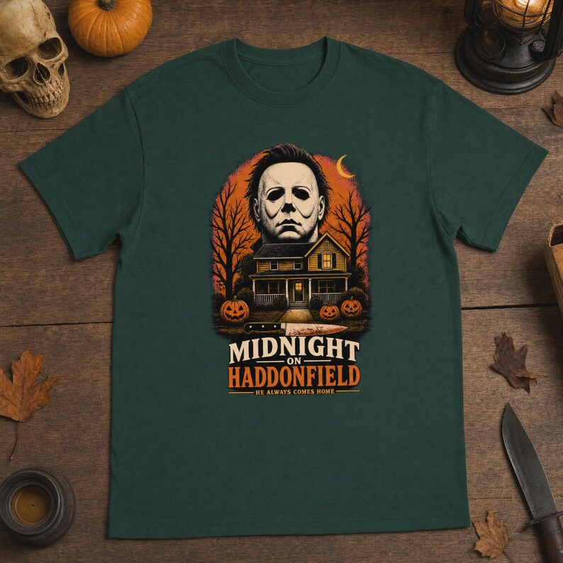 May include: A dark green t-shirt featuring a graphic print of a masked figure above a two-story house, trees, and pumpkins. The text reads MIDNIGHT ON HADDONFIELD HE ALWAYS COMES HOME. A knife is illustrated horizontally below the house.