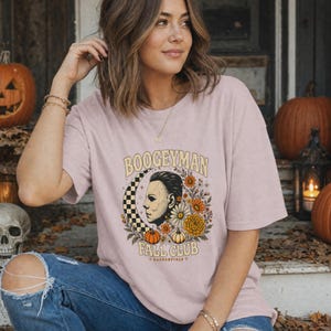 May include: A light pink short-sleeve t-shirt featuring a graphic print of a masked face, a checkered crescent moon, and various autumn flowers and pumpkins. The text reads BOOGEYMAN FALL CLUB and HADDONFIELD in a stylised font.