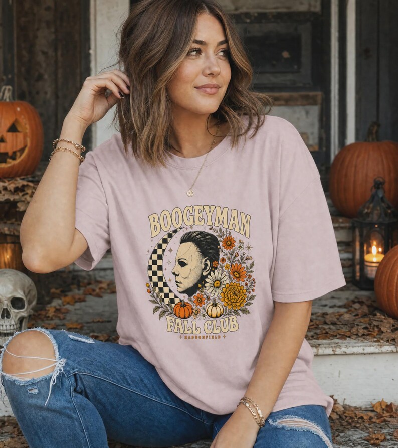 May include: A light pink short-sleeve t-shirt featuring a graphic print of a masked face, a checkered crescent moon, and various autumn flowers and pumpkins. The text reads BOOGEYMAN FALL CLUB and HADDONFIELD in a stylised font.