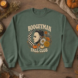 May include: A forest green crewneck sweatshirt featuring a graphic print of a masked figure in profile, a checkered crescent moon, orange and white flowers, and small pumpkins. The text reads BOOGEYMAN FALL CLUB in a curved cream-coloured font.