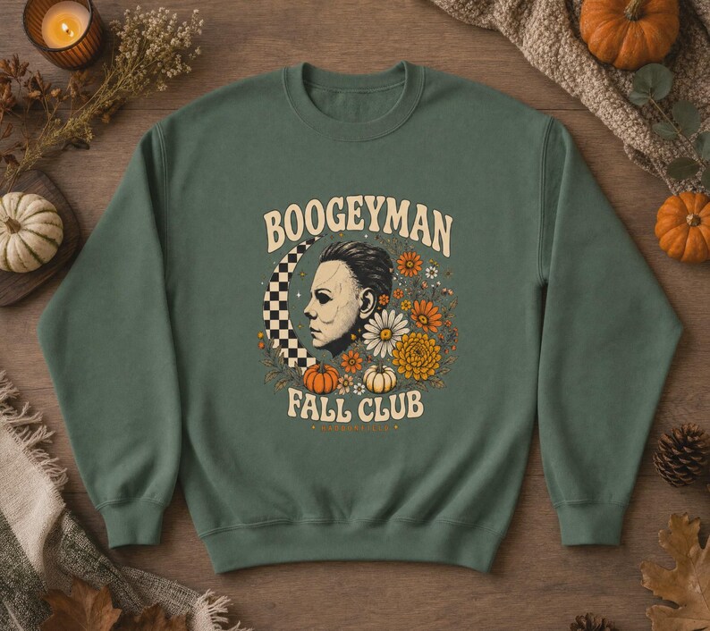 May include: A forest green crewneck sweatshirt featuring a graphic print of a masked figure in profile, a checkered crescent moon, orange and white flowers, and small pumpkins. The text reads BOOGEYMAN FALL CLUB in a curved cream-coloured font.