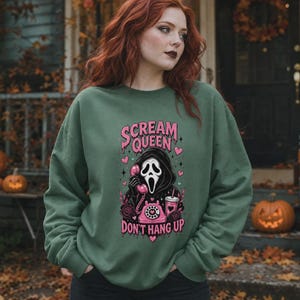 May include: An olive green crewneck sweatshirt featuring a graphic print of a masked figure holding a pink rotary telephone. The text reads "SCREAM QUEEN" and "DON'T HANG UP" in pink lettering, surrounded by pink hearts, roses, and a drink cup.