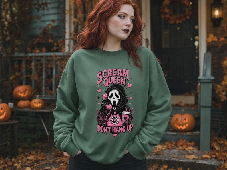 May include: An olive green crewneck sweatshirt featuring a graphic print of a masked figure holding a pink rotary telephone. The text reads "SCREAM QUEEN" and "DON'T HANG UP" in pink lettering, surrounded by pink hearts, roses, and a drink cup.