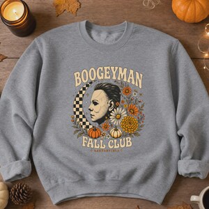 May include: A heather grey crewneck sweatshirt featuring a graphic print of a masked figure in profile, a checkered crescent moon, orange and white flowers, and pumpkins. The text reads BOOGEYMAN FALL CLUB in a curved, light-coloured font.