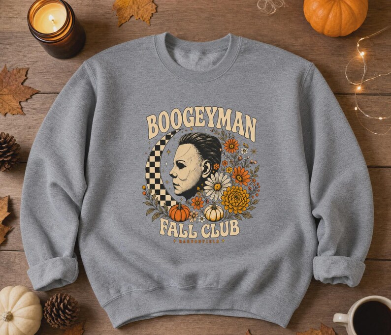 May include: A heather grey crewneck sweatshirt featuring a graphic print of a masked figure in profile, a checkered crescent moon, orange and white flowers, and pumpkins. The text reads BOOGEYMAN FALL CLUB in a curved, light-coloured font.