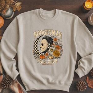 May include: A beige crewneck sweatshirt featuring a graphic print of a profile of a man, a checkered crescent moon, orange and white pumpkins, and various autumn flowers. The text reads BOOGEYMAN FALL CLUB and HADDONFIELD in a retro font.