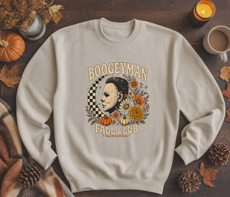 May include: A beige crewneck sweatshirt featuring a graphic print of a profile of a man, a checkered crescent moon, orange and white pumpkins, and various autumn flowers. The text reads BOOGEYMAN FALL CLUB and HADDONFIELD in a retro font.