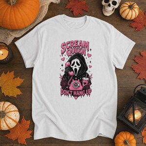 May include: A white t-shirt featuring a graphic of a hooded figure with a white mask holding a pink rotary telephone. The text reads "SCREAM QUEEN" and "DON'T HANG UP" in pink, with pink hearts, stars, and a pink drink cup surrounding the figure.