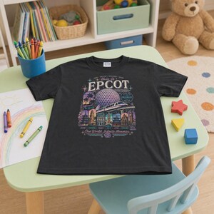May include: A black short-sleeved t-shirt featuring a graphic print of the Epcot Spaceship Earth structure, a monorail, and various international pavilion silhouettes with the text "This is Epcot" and "One World, Infinite Memories" in a colourful design.