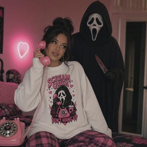 May include: A white long-sleeve sweatshirt featuring a graphic print of a masked figure holding a pink rotary phone, surrounded by pink hearts and stars. The text reads "SCREAM QUEEN" at the top and "DON'T HANG UP" at the bottom in pink lettering.