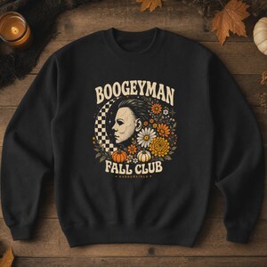 May include: A black crewneck sweatshirt featuring a graphic print of a masked face in profile, surrounded by flowers, pumpkins, and a checkered crescent moon. The text reads BOOGEYMAN FALL CLUB and HADDONFIELD in a cream-coloured retro font.