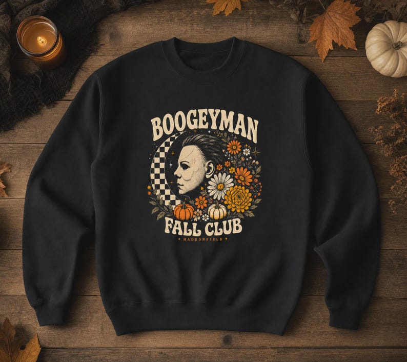 May include: A black crewneck sweatshirt featuring a graphic print of a masked face in profile, surrounded by flowers, pumpkins, and a checkered crescent moon. The text reads BOOGEYMAN FALL CLUB and HADDONFIELD in a cream-coloured retro font.