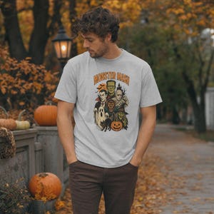 May include: A light gray short-sleeve t-shirt featuring a graphic print of classic Halloween monsters including a vampire, mummy, werewolf, and witch. The text "MONSTER MASH" is printed in orange above the illustration of the spooky characters.