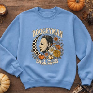 May include: A light blue crewneck sweatshirt featuring a graphic print with the text BOOGEYMAN FALL CLUB. The design includes a profile of a masked figure, a checkered crescent moon, orange and white pumpkins, and various autumn-coloured flowers.