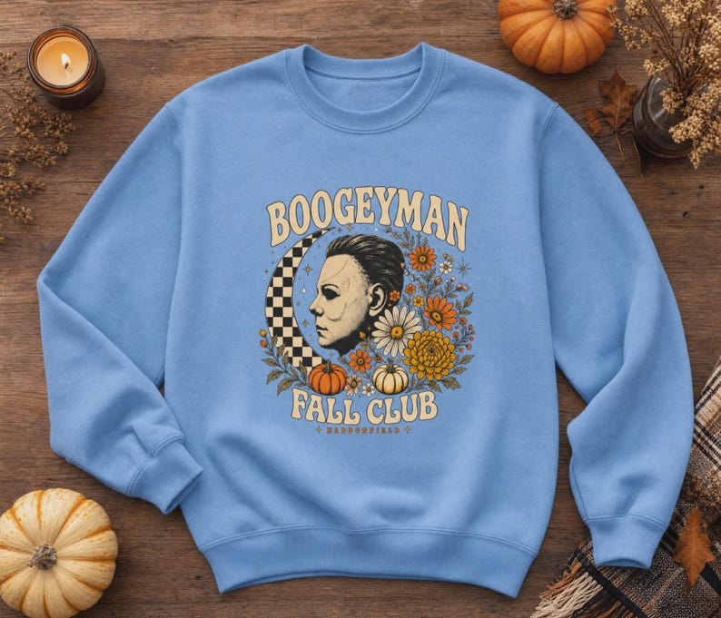 May include: A light blue crewneck sweatshirt featuring a graphic print with the text BOOGEYMAN FALL CLUB. The design includes a profile of a masked figure, a checkered crescent moon, orange and white pumpkins, and various autumn-coloured flowers.