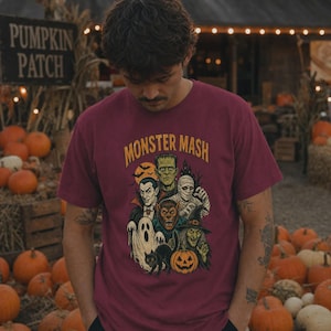 May include: A maroon short-sleeve t-shirt featuring a graphic print with the text "MONSTER MASH" above illustrations of Frankenstein's monster, a vampire, a mummy, a werewolf, a ghost, a witch, a black cat, and a jack-o'-lantern.