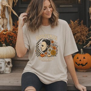 May include: A beige short-sleeve t-shirt featuring a graphic print with the text BOOGEYMAN FALL CLUB HADDONFIELD. The design includes a profile illustration of a masked figure, a checkered crescent moon, orange and yellow flowers, and pumpkins.