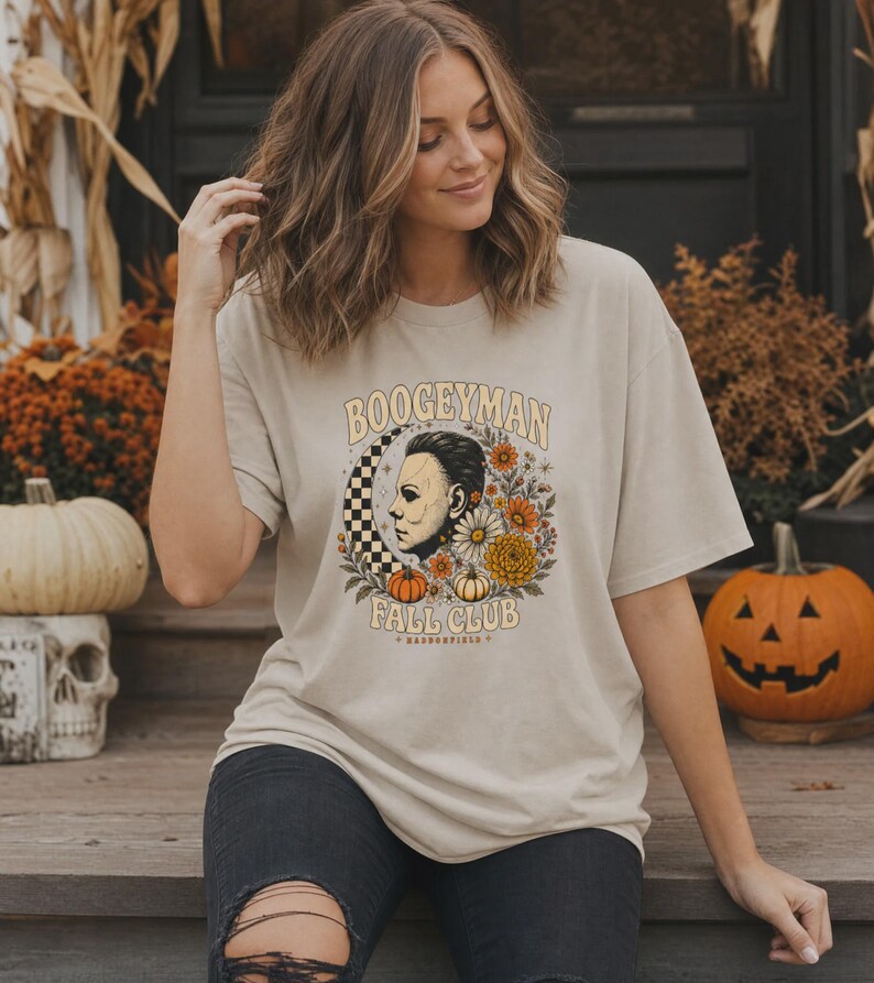 May include: A beige short-sleeve t-shirt featuring a graphic print with the text BOOGEYMAN FALL CLUB HADDONFIELD. The design includes a profile illustration of a masked figure, a checkered crescent moon, orange and yellow flowers, and pumpkins.