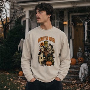 May include: A beige crewneck sweatshirt featuring a graphic print with the text Monster Mash. The design displays a collection of classic Halloween characters including a vampire, Frankenstein's monster, a mummy, a werewolf, a witch, and a pumpkin.