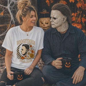 May include: A white short-sleeved t-shirt featuring a graphic print with the text BOOGEYMAN FALL CLUB, a profile illustration of a masked figure, a checkered crescent moon, and autumn flowers. Two black mugs with orange jack-o'-lantern faces.