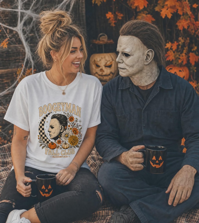 May include: A white short-sleeved t-shirt featuring a graphic print with the text BOOGEYMAN FALL CLUB, a profile illustration of a masked figure, a checkered crescent moon, and autumn flowers. Two black mugs with orange jack-o'-lantern faces.