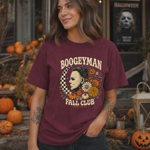 May include: A maroon short-sleeved t-shirt featuring a graphic print of a masked figure in profile, a checkered crescent moon, pumpkins, and flowers. The text reads "BOOGEYMAN FALL CLUB" in a cream-coloured font, with "HADDONFIELD" below.