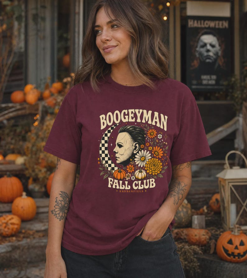 May include: A maroon short-sleeved t-shirt featuring a graphic print of a masked figure in profile, a checkered crescent moon, pumpkins, and flowers. The text reads "BOOGEYMAN FALL CLUB" in a cream-coloured font, with "HADDONFIELD" below.
