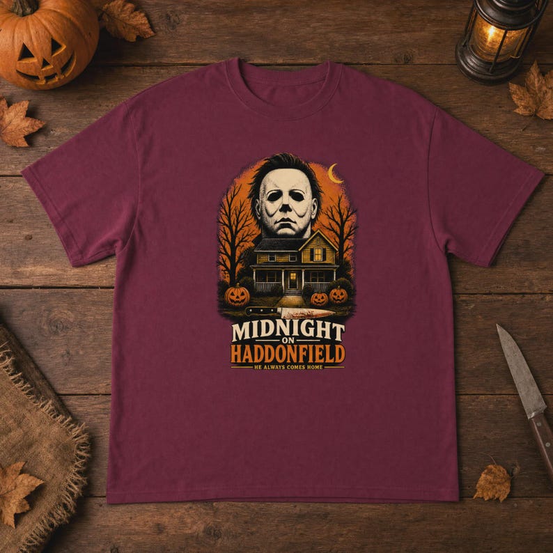 May include: A maroon short-sleeve t-shirt featuring a graphic of a masked face, a house, and pumpkins. The text reads MIDNIGHT ON HADDONFIELD HE ALWAYS COMES HOME. The design includes a knife illustration and a crescent moon against a dark orange background.