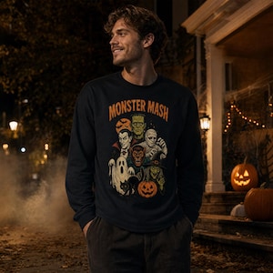 May include: A black long-sleeve sweatshirt featuring a graphic print with the text MONSTER MASH in orange letters. The design includes illustrations of a vampire, Frankenstein's monster, a mummy, a werewolf, a ghost, a witch, and a pumpkin.