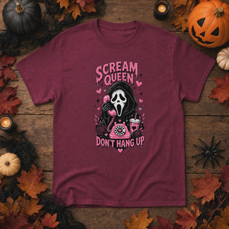 May include: A maroon t-shirt featuring a graphic of a hooded figure in a white mask holding a pink rotary telephone. The text reads "SCREAM QUEEN" and "DON'T HANG UP" in pink lettering, surrounded by pink hearts, roses, and a pink drink cup.