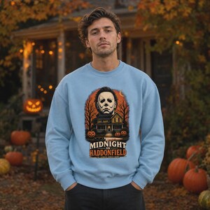 May include: A light blue crewneck sweatshirt featuring a graphic print of a masked figure above a house, surrounded by bare trees and pumpkins. The text reads "MIDNIGHT ON HADDONFIELD" and "HE ALWAYS COMES HOME" with a knife illustration below.