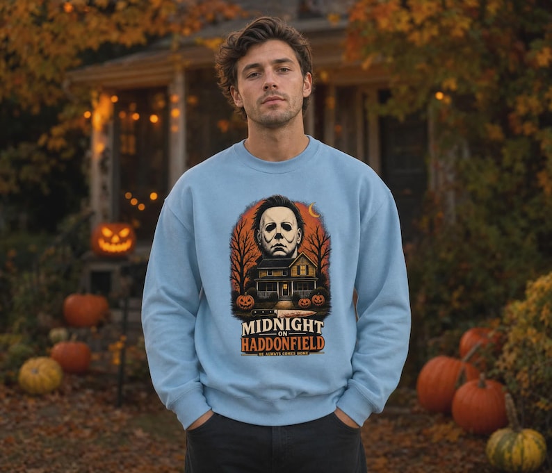 May include: A light blue crewneck sweatshirt featuring a graphic print of a masked figure above a house, surrounded by bare trees and pumpkins. The text reads "MIDNIGHT ON HADDONFIELD" and "HE ALWAYS COMES HOME" with a knife illustration below.