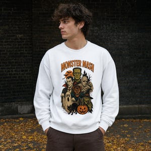 May include: A white crewneck sweatshirt featuring a colorful graphic print of classic Halloween monsters including a vampire, mummy, werewolf, ghost, witch, and jack-o'-lantern, with the orange text Monster Mash arched above the illustration.