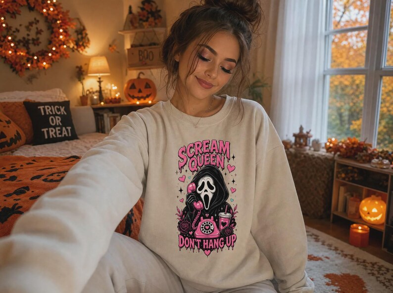 May include: A beige crewneck sweatshirt featuring a graphic print of a hooded figure in a mask holding a pink rotary telephone. The text reads Scream Queen Don't Hang Up in pink lettering, surrounded by small pink hearts and floral accents.