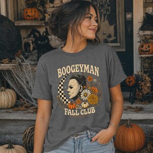 May include: A dark grey short-sleeve t-shirt featuring a graphic print with the text BOOGEYMAN FALL CLUB. The design includes a profile illustration of a masked figure, a chequered crescent moon, orange and white flowers, and small pumpkins.