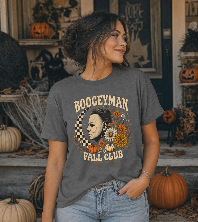 May include: A dark grey short-sleeve t-shirt featuring a graphic print with the text BOOGEYMAN FALL CLUB. The design includes a profile illustration of a masked figure, a chequered crescent moon, orange and white flowers, and small pumpkins.