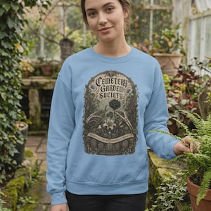 May include: A light blue crewneck sweatshirt featuring a graphic print of the text Cemetery Garden Society, skeletal hands, a black rose, and gravestones. The design is framed in an ornate archway with botanical details and moths.