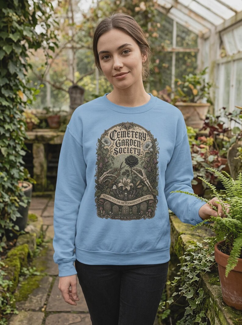 May include: A light blue crewneck sweatshirt featuring a graphic print of the text Cemetery Garden Society, skeletal hands, a black rose, and gravestones. The design is framed in an ornate archway with botanical details and moths.