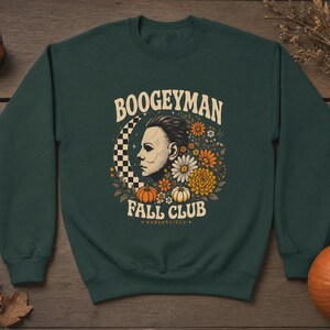 May include: A dark forest green crewneck sweatshirt featuring a graphic print with the text BOOGEYMAN FALL CLUB HADDONFIELD. The design includes a profile of a white mask, a checkered crescent moon, orange pumpkins, and various autumn flowers.