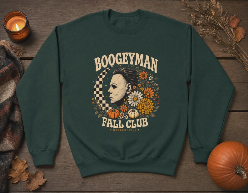 May include: A dark forest green crewneck sweatshirt featuring a graphic print with the text BOOGEYMAN FALL CLUB HADDONFIELD. The design includes a profile of a white mask, a checkered crescent moon, orange pumpkins, and various autumn flowers.