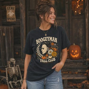 May include: A charcoal grey short-sleeve t-shirt featuring a graphic print with the text "BOOGEYMAN FALL CLUB" and "HADDONFIELD". The design includes a profile illustration of a masked figure, a checkered crescent moon, and orange flowers.